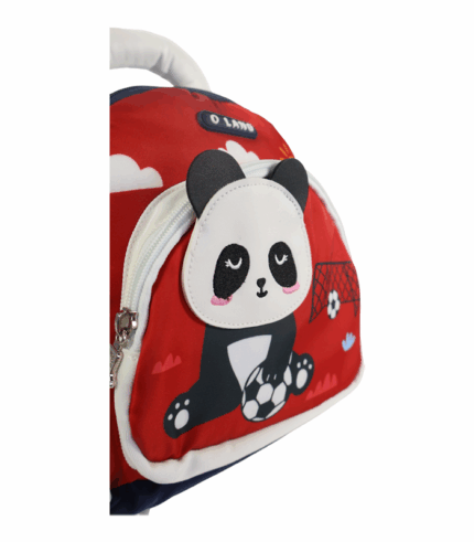 Detalle de una mochila infantil roja con cremallera blanca y un parche bordado de un panda sosteniendo un balón de fútbol sobre un fondo de portería y nubes.