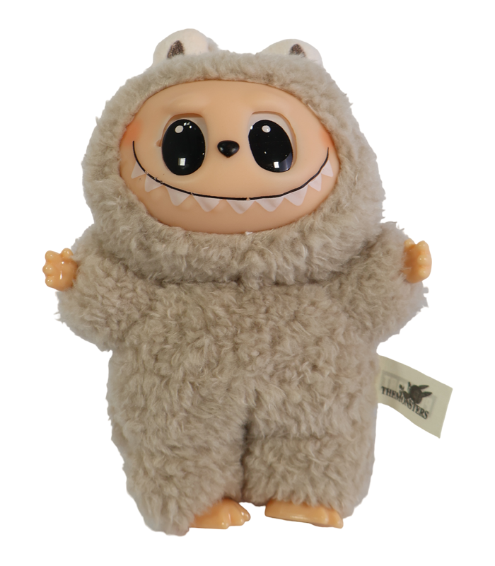 Primer plano de una figura coleccionable Labubu, un monstruo beige cubierto de peluche, con la cara, manos y pies de vinilo color piel, sonriendo ampliamente y con grandes ojos negros. Se aprecia la etiqueta de 'The Monsters' en el lateral.