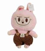 Peluche de personaje Labubu de color rosa pálido y textura suave. Viste unos overoles de pana marrón oscuro con tirantes y pequeños parches decorativos en forma de osos y una etiqueta que dice 'HAPPY'. El muñeco tiene orejas de conejo y una expresión sonriente y pícara con dientes bordados.
